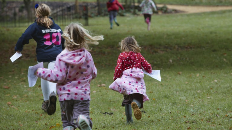 Children in waterproof clothing running across a lawn away from the camera, clutching trail sheets